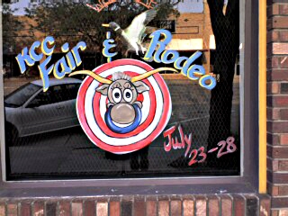 [KCC Fair & Rodeo written about a bull coming out of a white and red-ringed target. The bull has a huge, blue nose ring. The dates July 23-28 are in the lower corner. This is painted on a window of a main street business.]